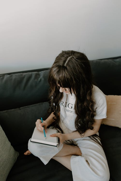 A Girl Writing in a Notebook while Sitting on a Sofa