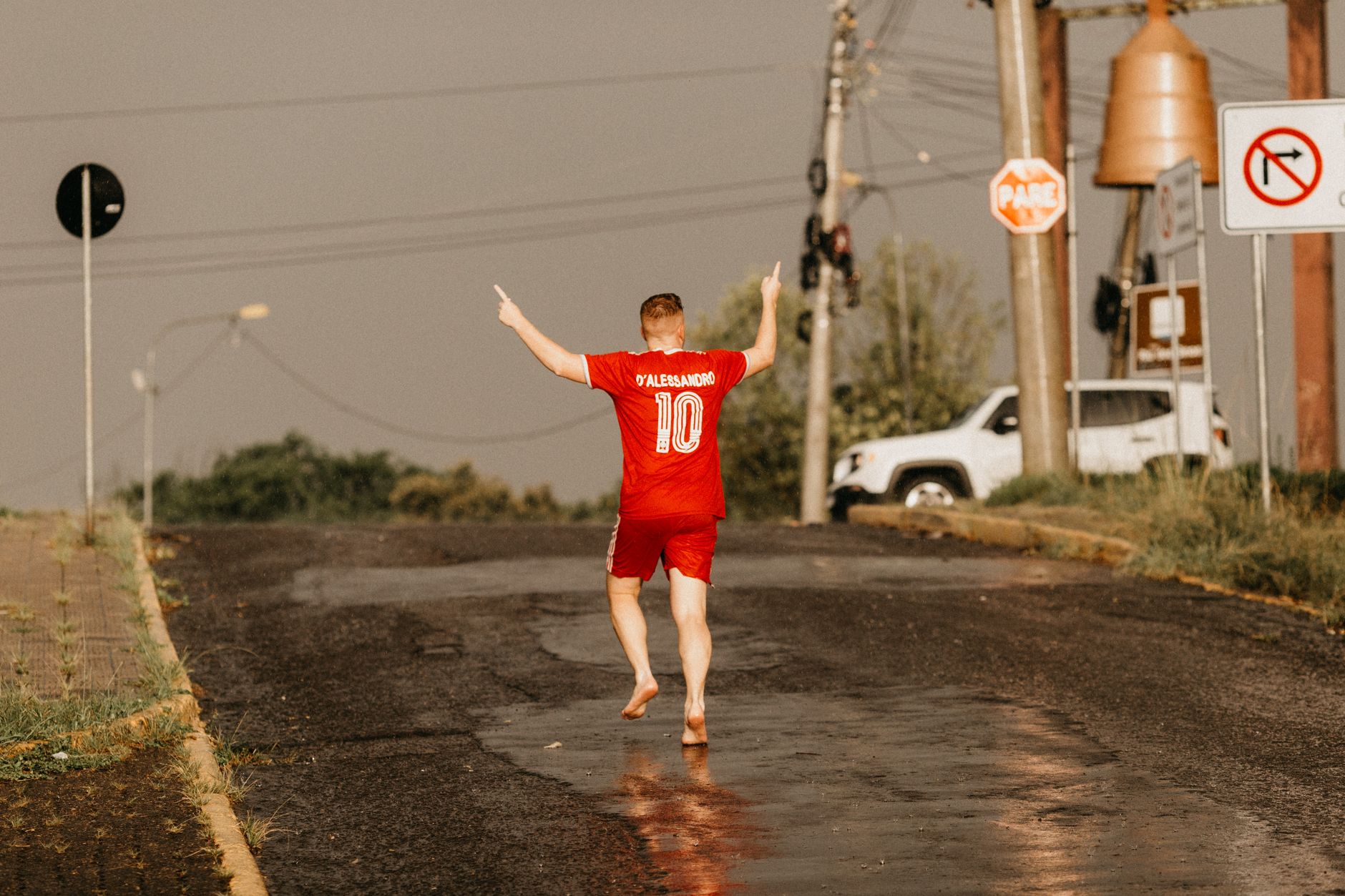 Man walking barefoot in football kit on wet urban street after rain, hands raised in celebration.