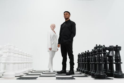 Two individuals stand on a giant chessboard surrounded by oversized chess pieces, symbolizing a mind game.