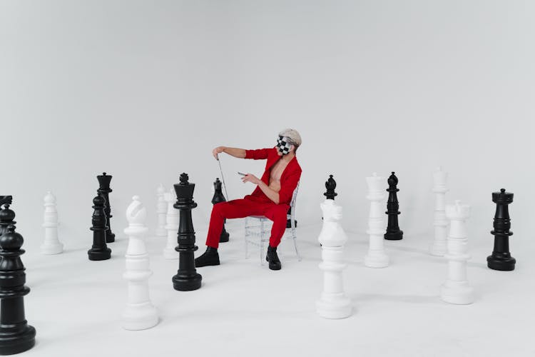 A Man In A Red Suit With Face Makeup Sitting Beside Giant Chess Pieces