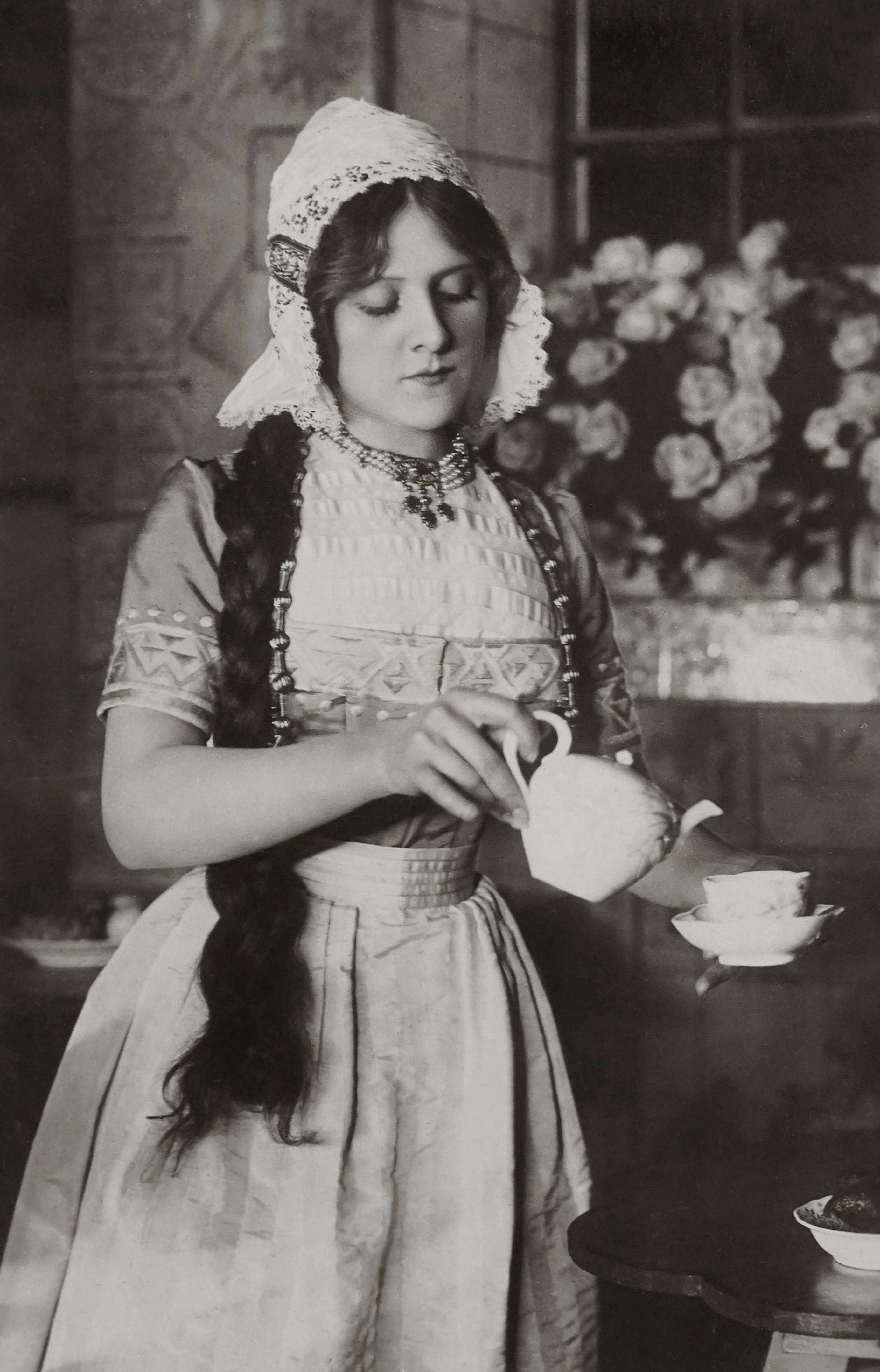 A woman in traditional attire pouring tea from a teapot in a vintage setting.