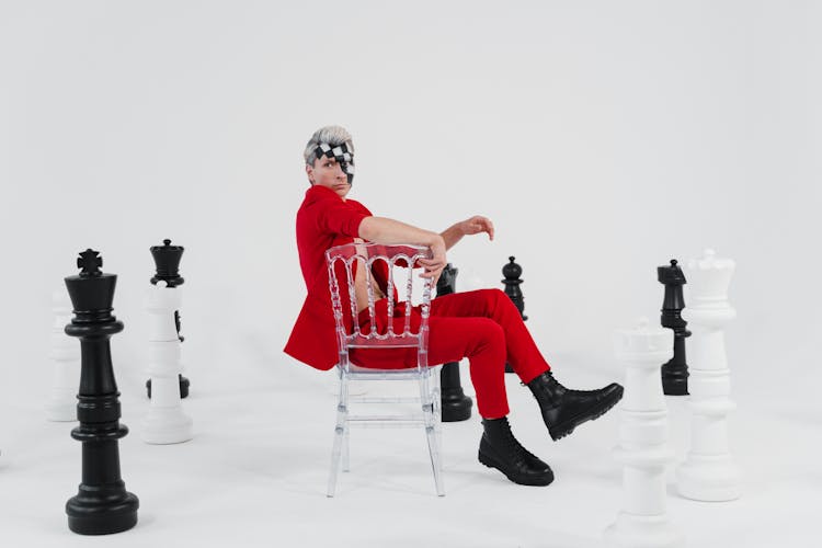 A Man In A Red Suit With Face Makeup Sitting Beside Giant Chess Pieces