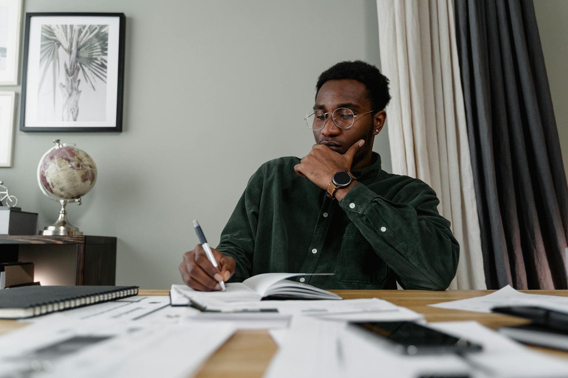 Black man in glasses concentrating over documents at a wooden desk, office setting.