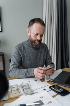 Bearded man in gray shirt counting dollar bills at office desk.