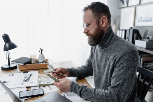 Businessman wearing glasses counting cash at office desk with documents and smartphone.