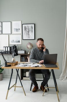 A man working in a modern office, multitasking with a phone and laptop at a wooden desk.