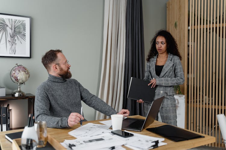 A Man In Gray Sweater Talking To His Employee In Plaid Blazer Holding Documents