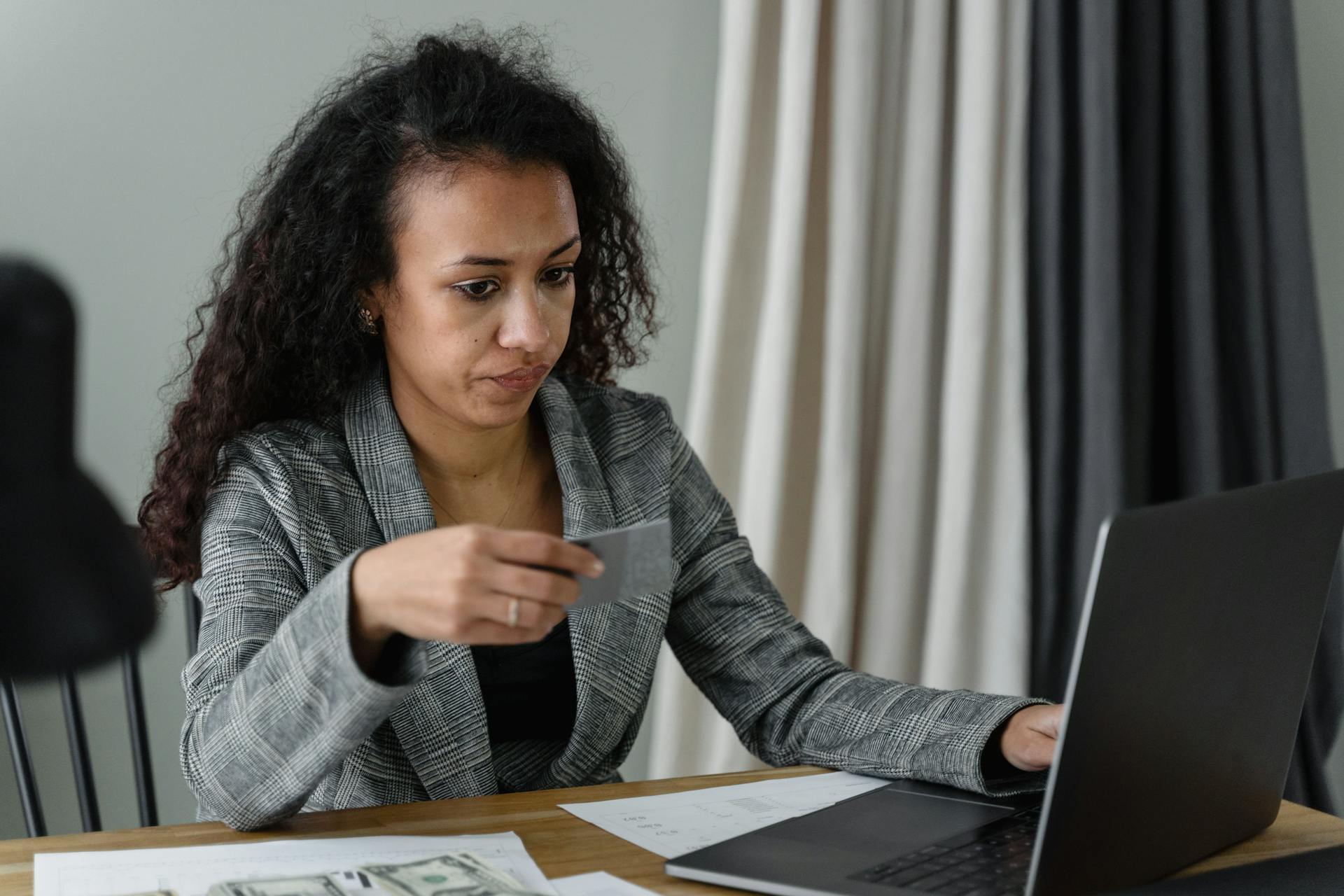 Professional woman at desk with credit card, laptop, and cash. Expression of concentration.