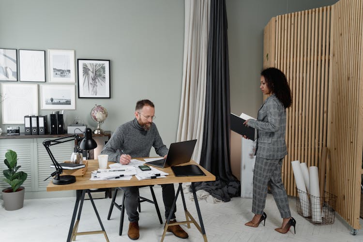 A Woman In Plaid Blazer And Pants Walking Towards The Man Sitting Near The Table With Laptop And Files