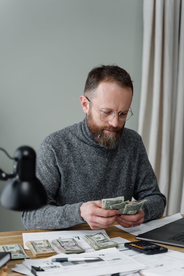 A Bearded Man In Gray Sweater Counting Dollars