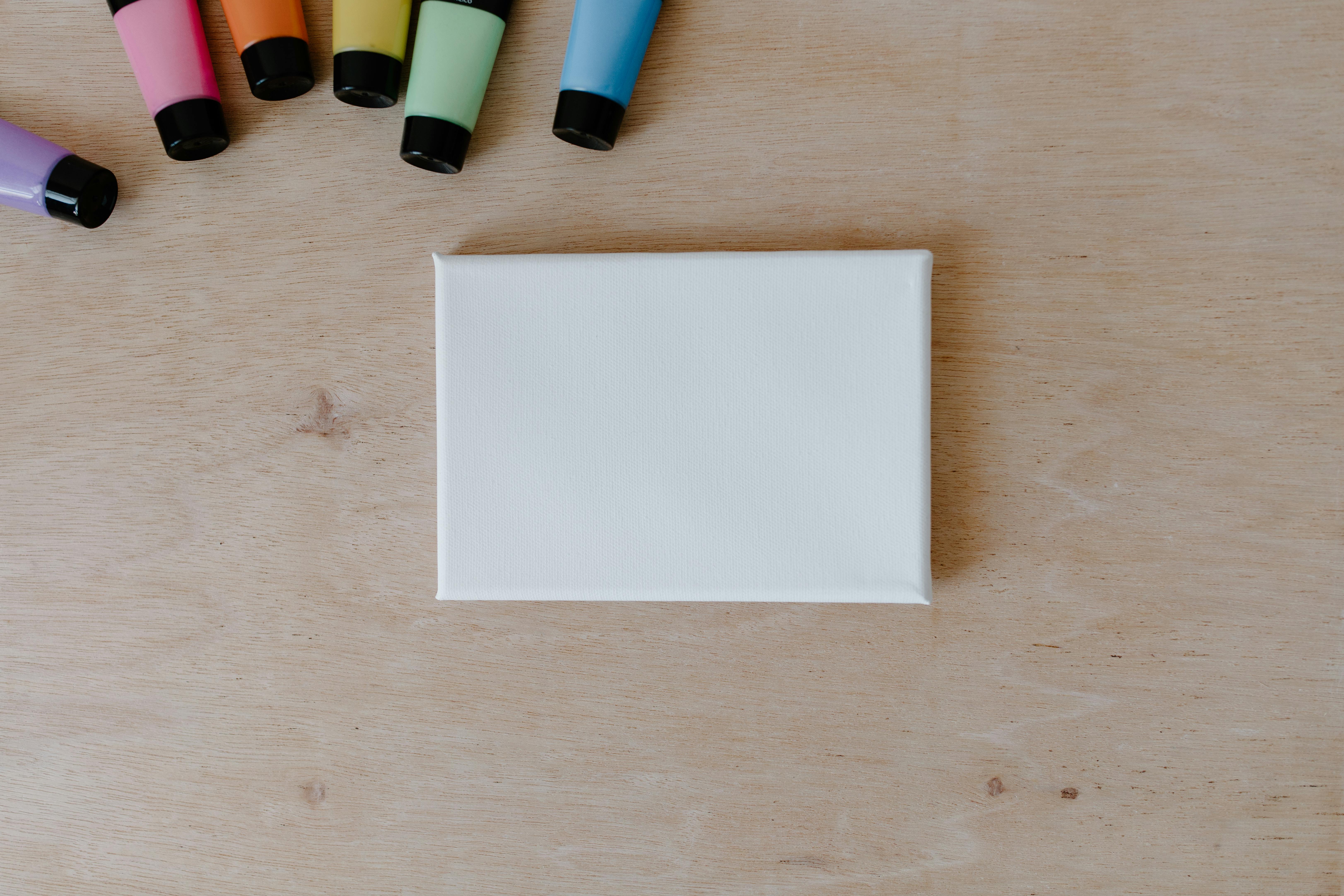 Top view of colorful paint tubes and blank canvas on a wooden surface.