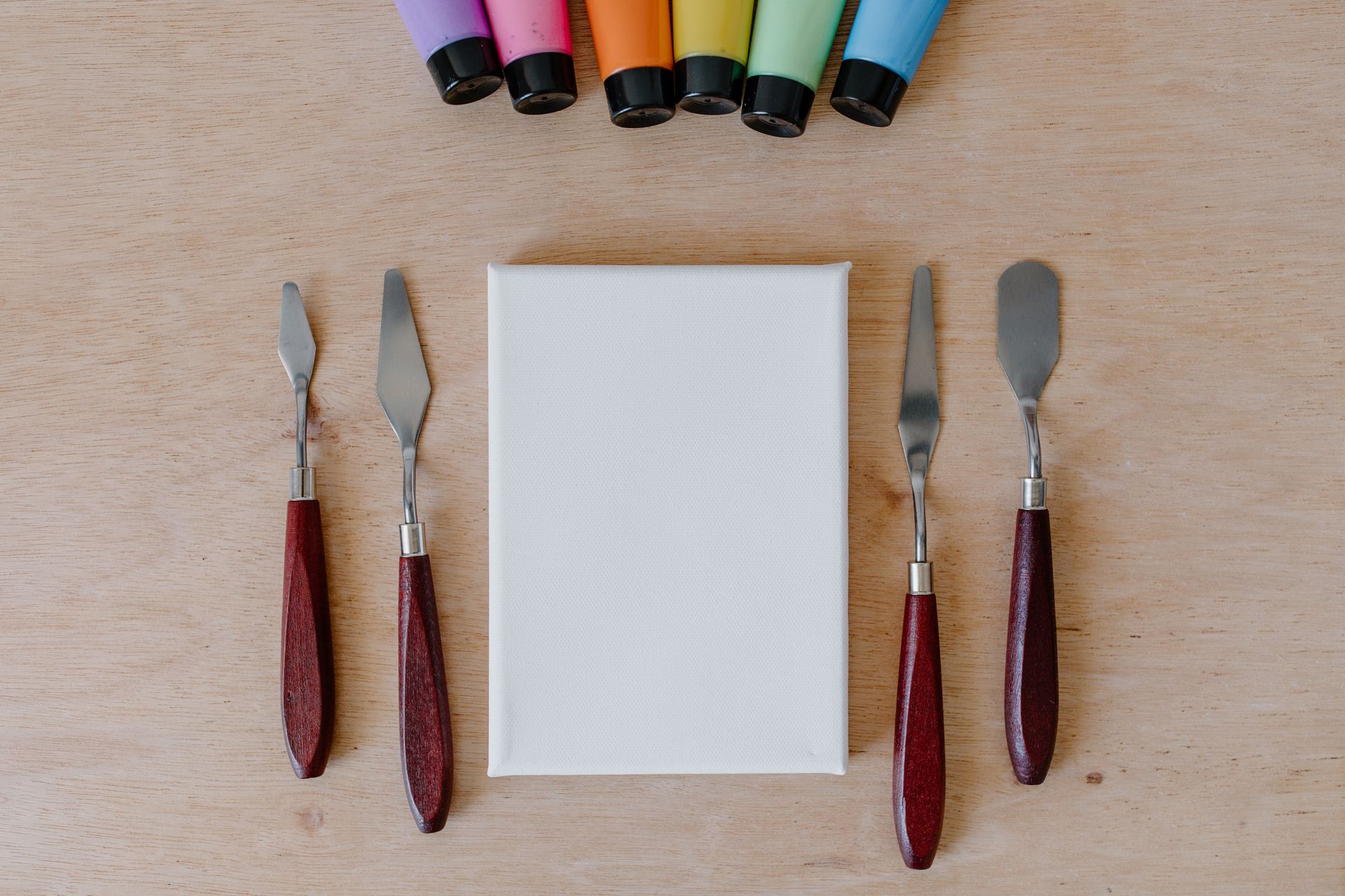 Flat lay of art supplies including canvas, paint, and palette knives on wooden surface.