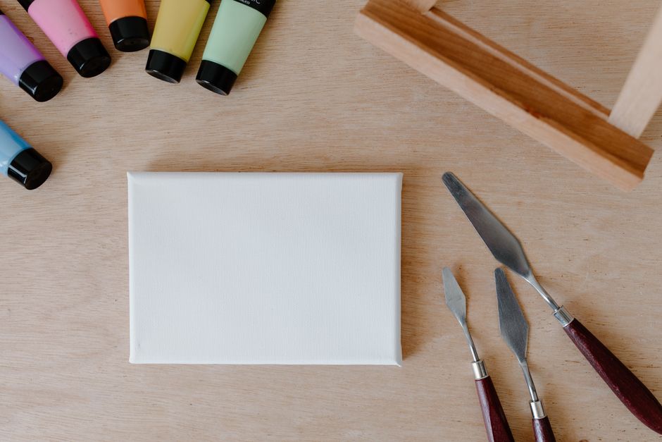 Top view of art supplies including paints, palette knives, and canvas on wooden surface.