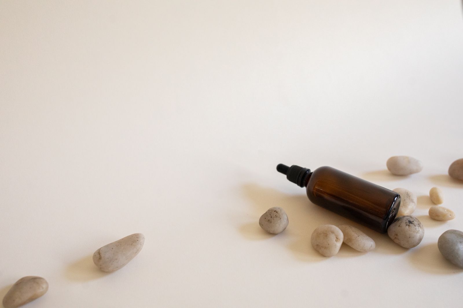 Amber glass dropper bottle lying among smooth pebbles on a warm white surface with soft shadows