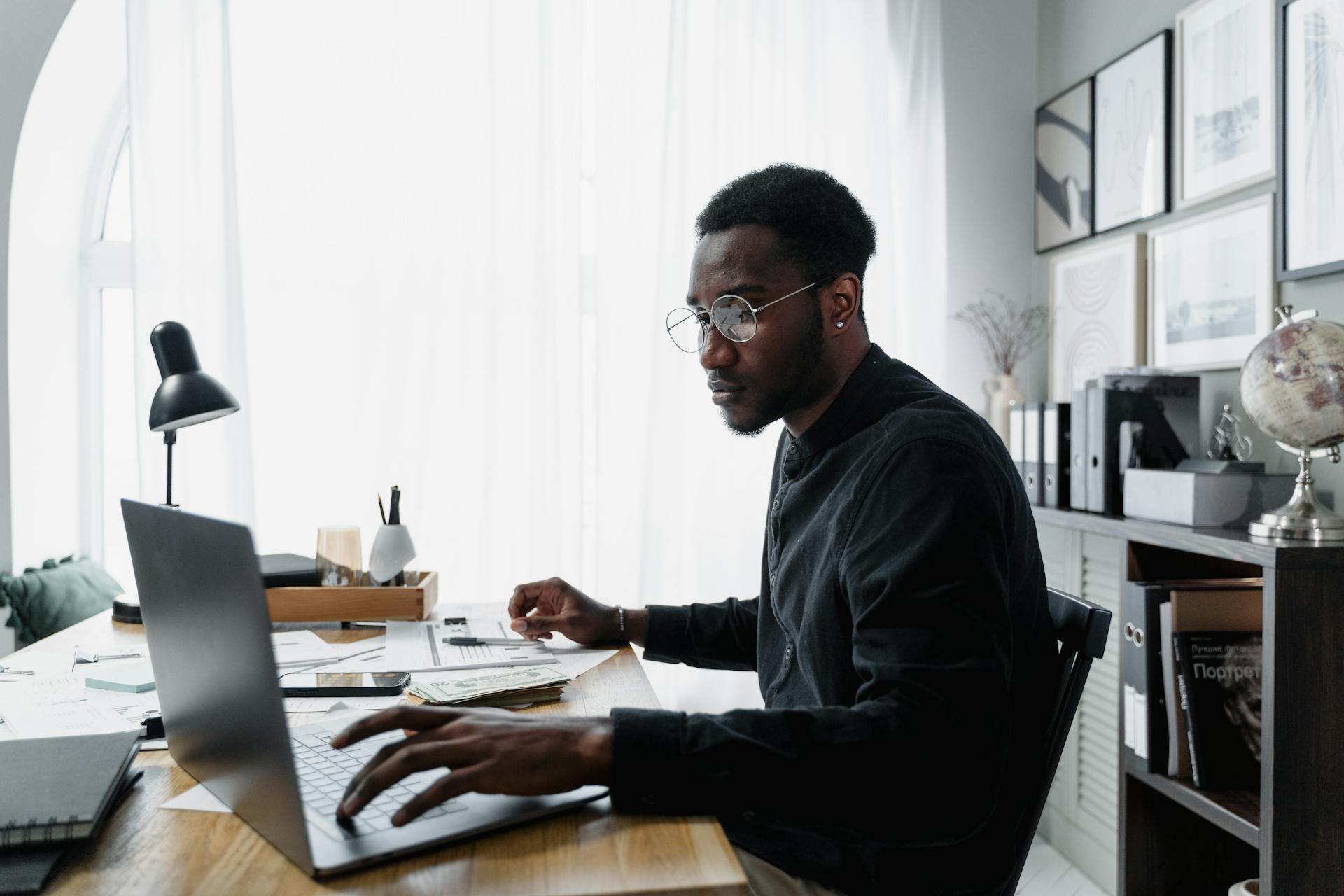 Black man in glasses working on laptop at office desk, surrounded by documents and modern decor.