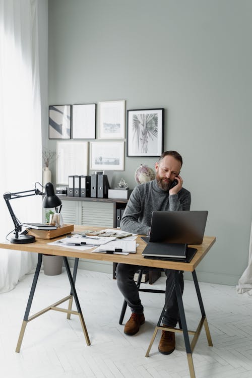 Free A Bearded Man Sitting at the Table with a Laptop Talking on the Phone Stock Photo