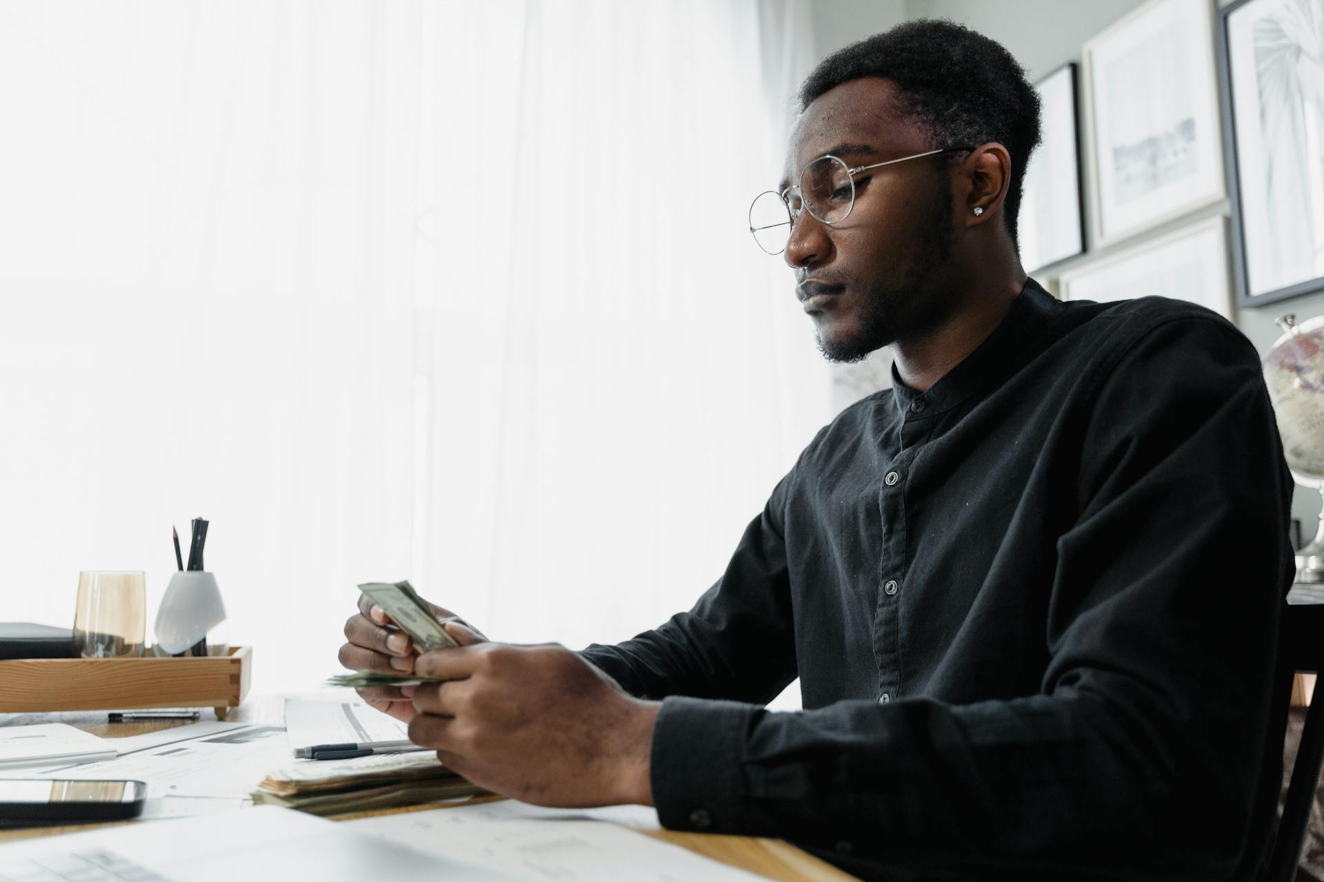 A focused man in glasses counts money while seated at a modern desk.