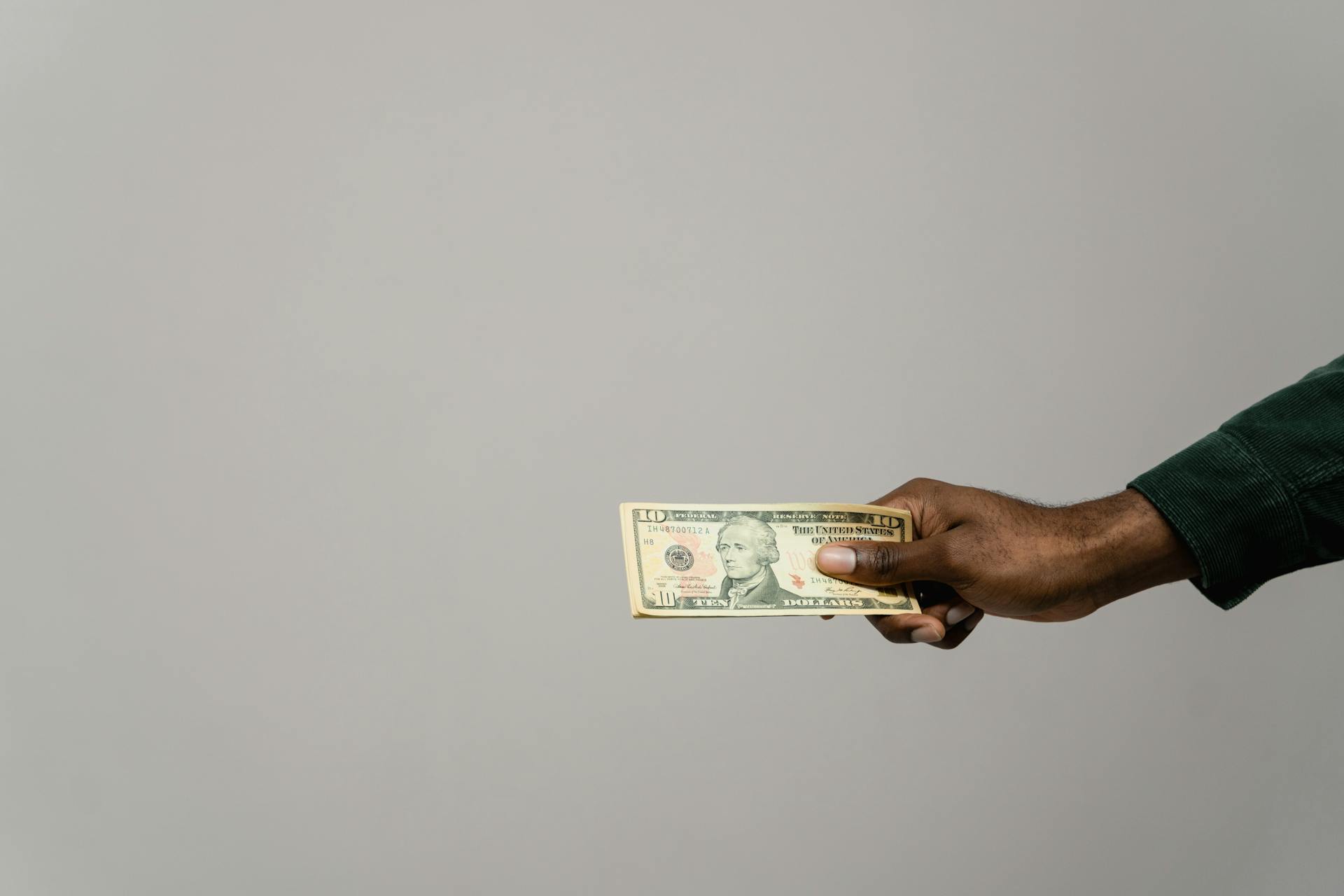 A hand extended holding a ten dollar bill against a neutral gray background, symbolizing payment or financial transaction.