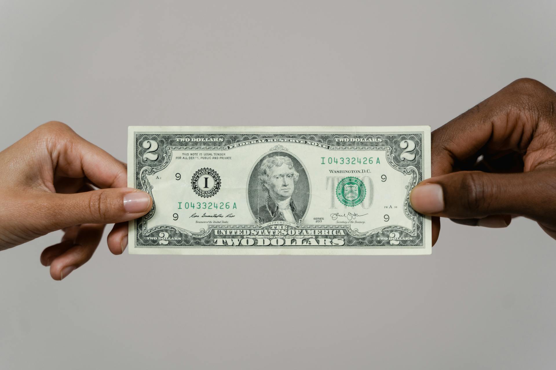 Close-up of two hands exchanging a two-dollar bill on a neutral background.