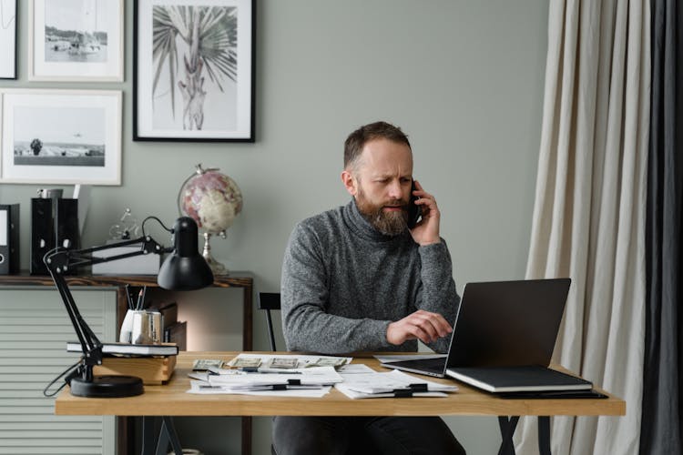 A Bearded Man Using A Laptop Near Cash Money On Wooden Table