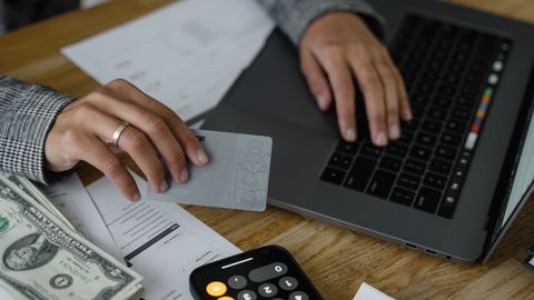 Close-up of a person using a credit card and laptop in a financial setting with cash and calculator.