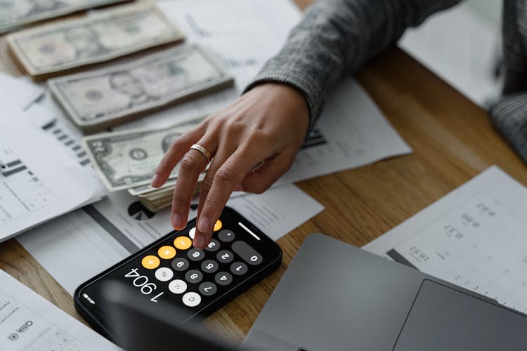 Hand Of A Person Using A Calculator Near Cash Money On Wooden Table