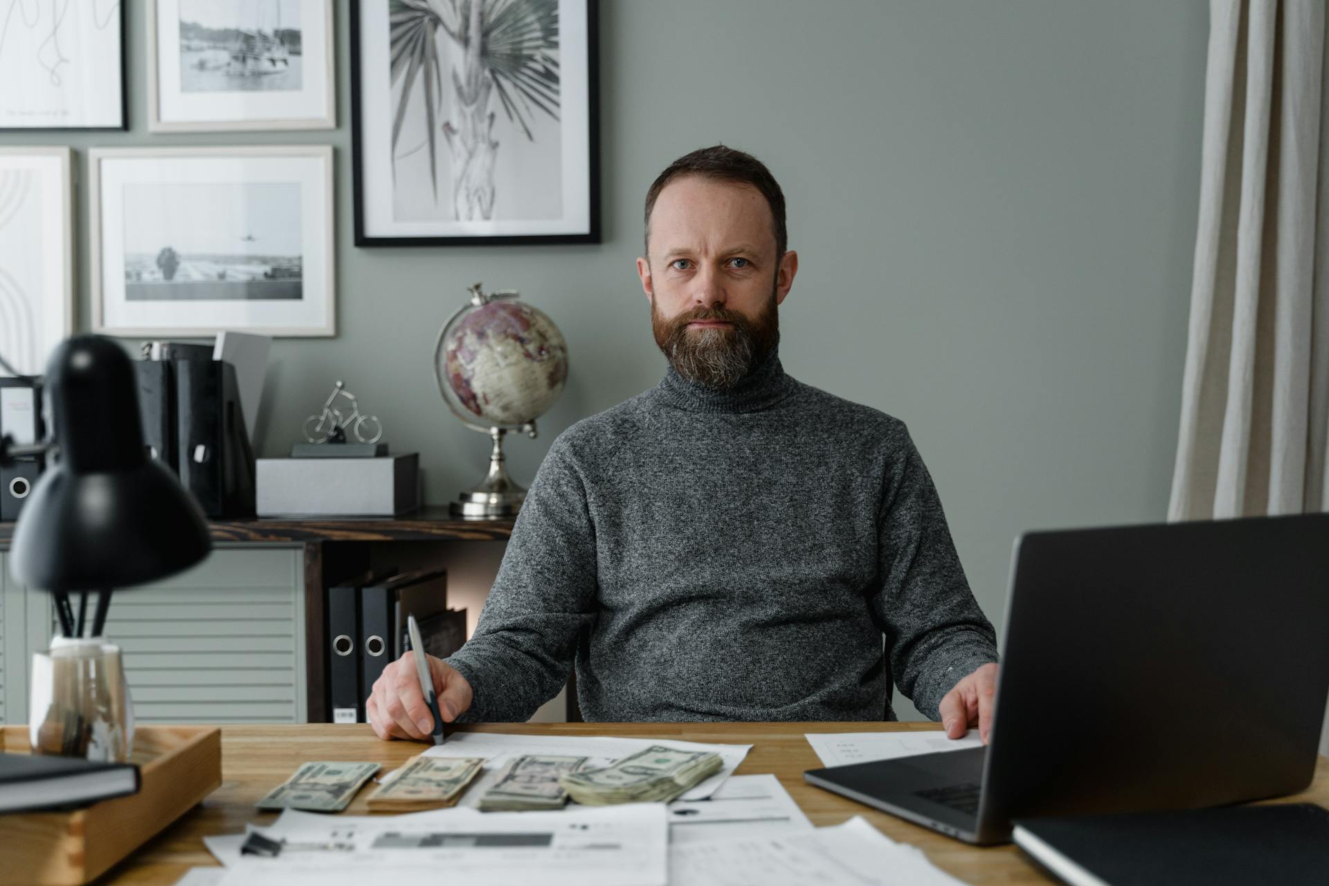 Bearded accountant in gray sweater working with banknotes and documents at office desk.