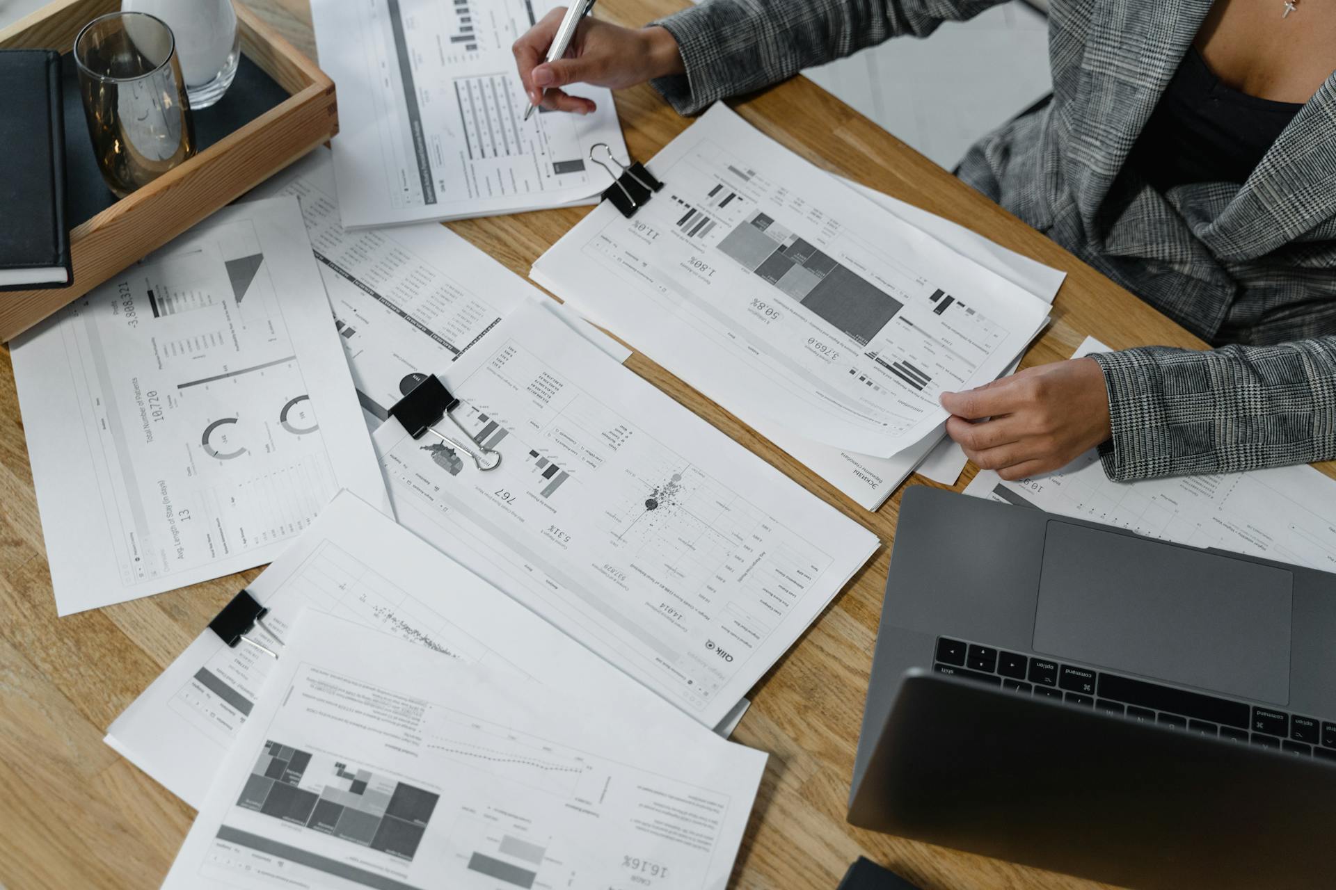 Woman analyzing financial data and reports on a wooden desk with a laptop, showcasing analytics in a workspace.