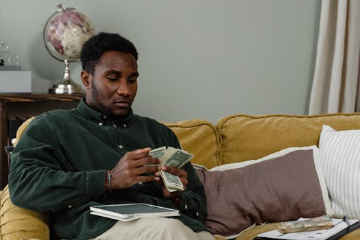 A man sitting on a couch counting cash notes in a cozy indoor setting with a globe in the background.