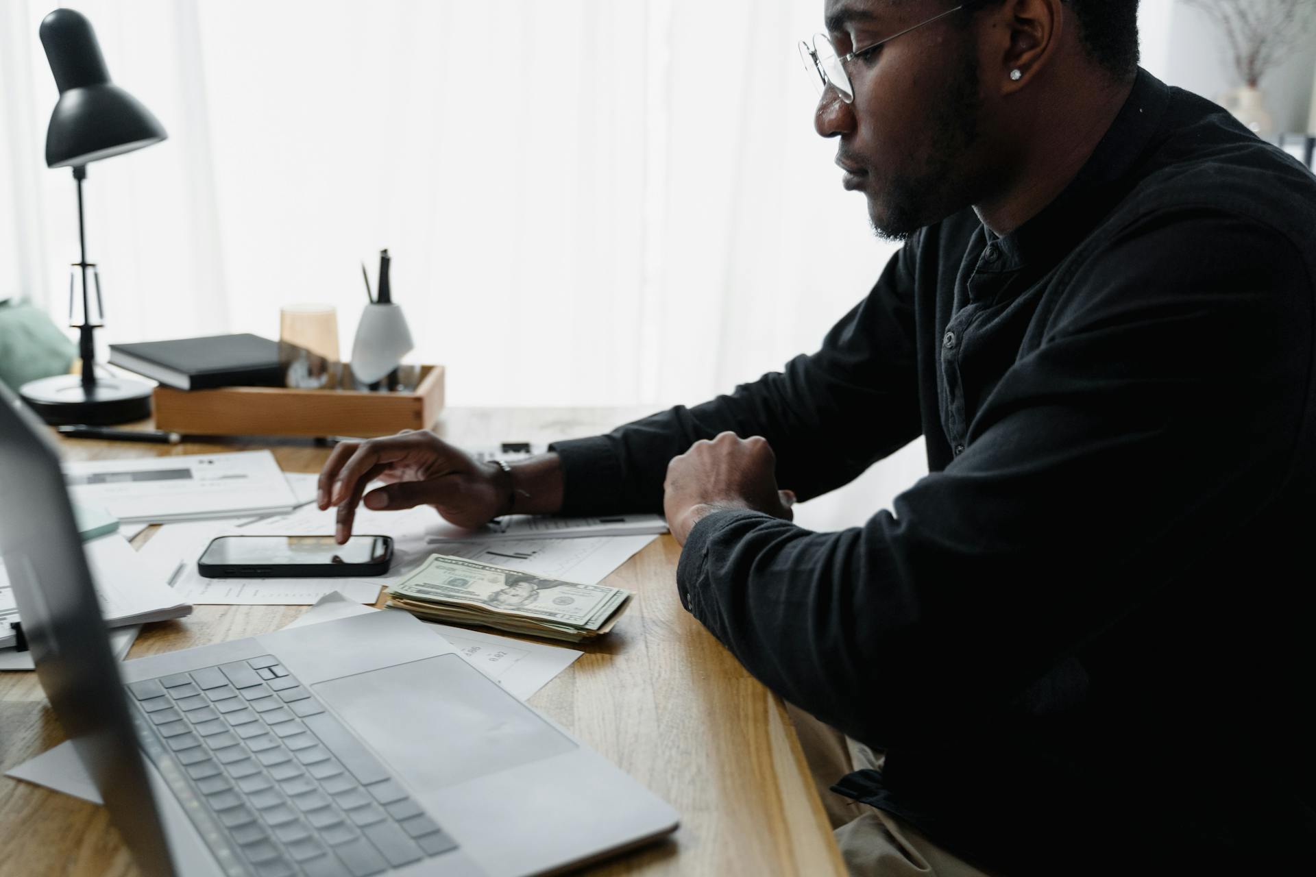 Businessman in office using smartphone for budgeting with cash and laptop on desk.