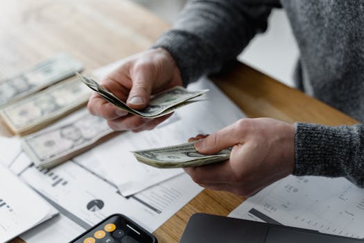 Person counting dollar bills on a desk with financial documents and a calculator in the background.