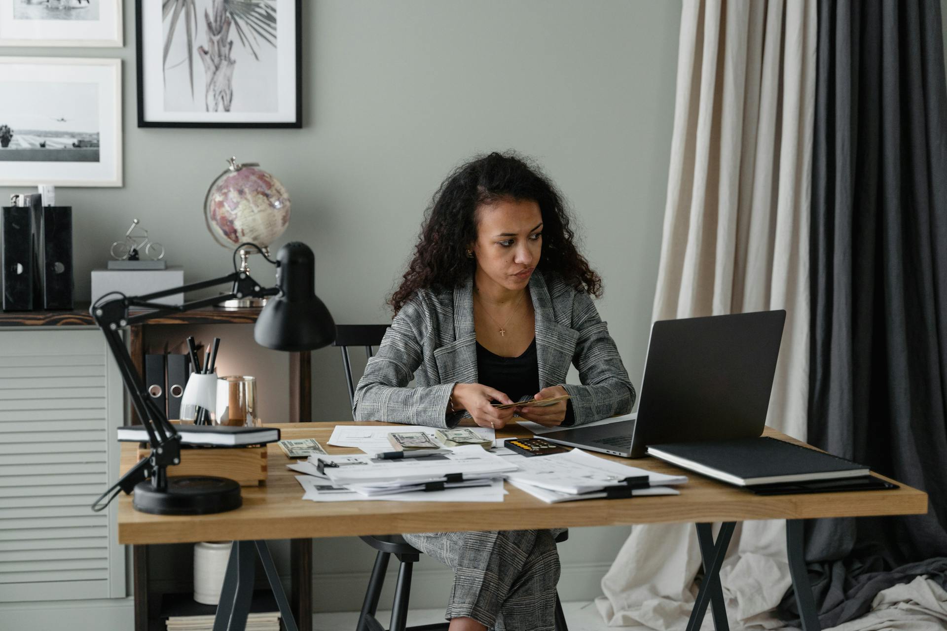 Woman in office reviewing finances with laptop, documents, and cash on table.