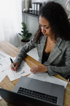 Businesswoman in office reviewing financial documents on a wooden desk with a laptop.