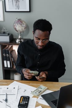 Adult African American man counting cash in an office setting with papers and a calculator.