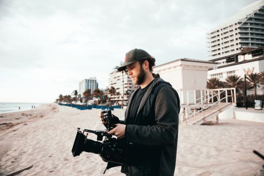 A filmmaker operates a camera on a sunny beachfront with modern buildings in view.