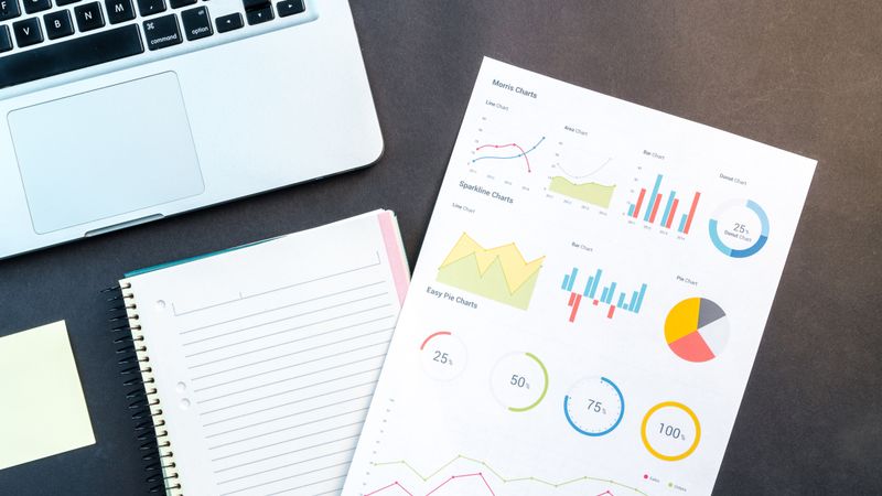 Flatlay of business report with colorful charts, a notebook, and a laptop for data analysis on a desk.