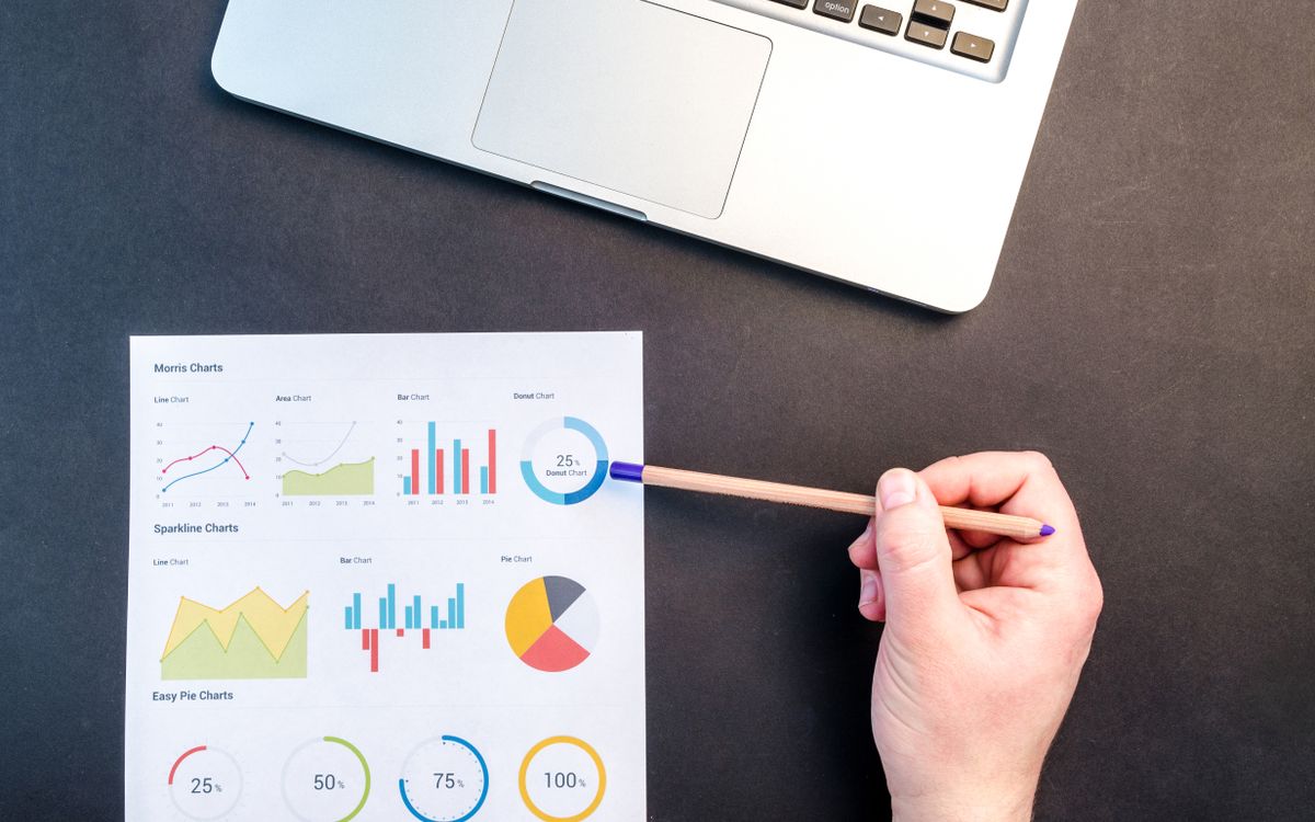 Hand holding pencil reviewing colorful data charts on desk with laptop.