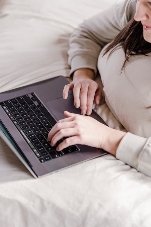 Free From above of unrecognizable female typing on netbook while lying under blanket on bed in bedroom during remote work at home Stock Photo