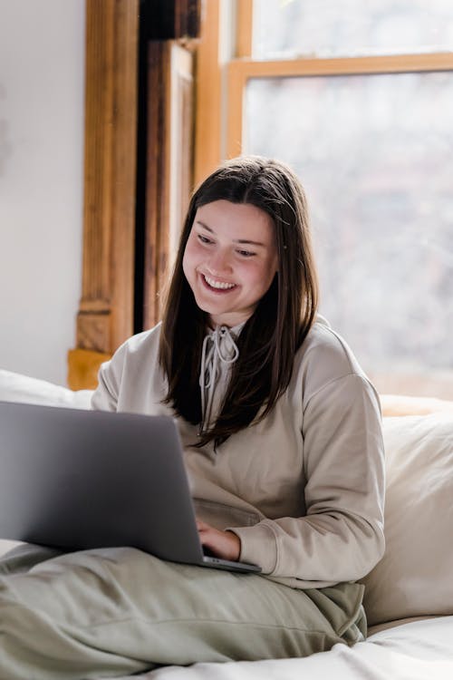 Free Happy female freelancer typing on netbook while sitting on comfortable bed during remote work at home in light cozy bedroom Stock Photo
