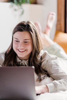 Young woman lying on bed, smiling while working on laptop in a cozy home setting.