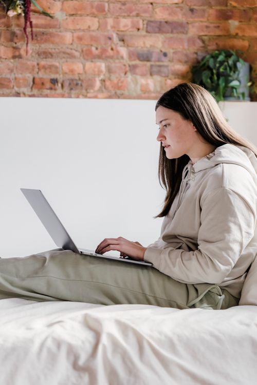 Free Side view of concentrated female freelancer typing on netbook while sitting on sofa bed near brick wall in light bedroom Stock Photo