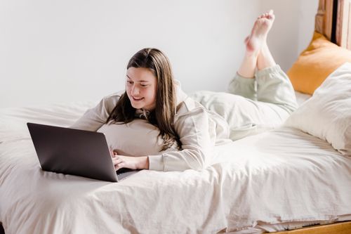 Free Full body of positive female freelancer typing on netbook while lying on comfortable white bed with raised legs during remote work Stock Photo