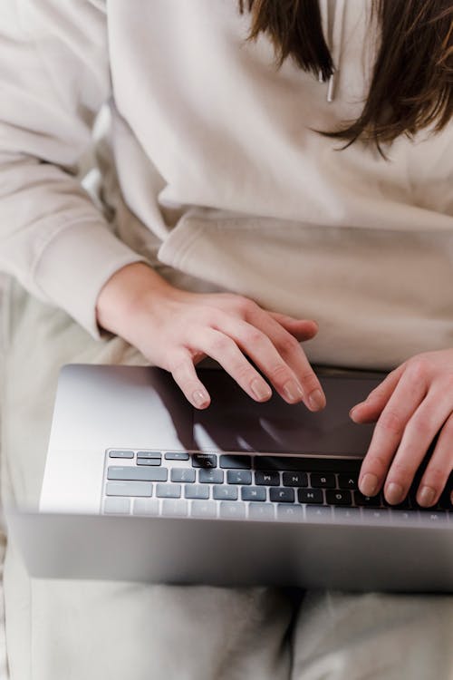 Unrecognizable female freelancer typing on keyboard of netbook while sitting under blanket in light bedroom during online work at home