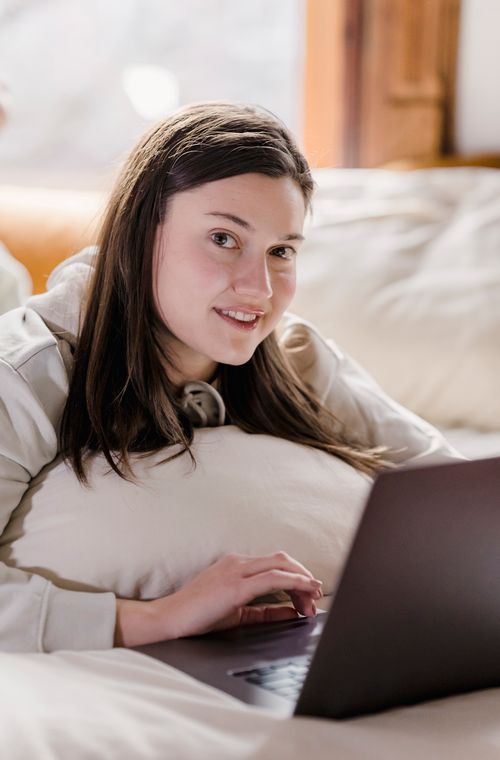 Free Positive female freelancer looking at camera and typing on modern netbook while lying on soft bed during remote work on blurred background Stock Photo