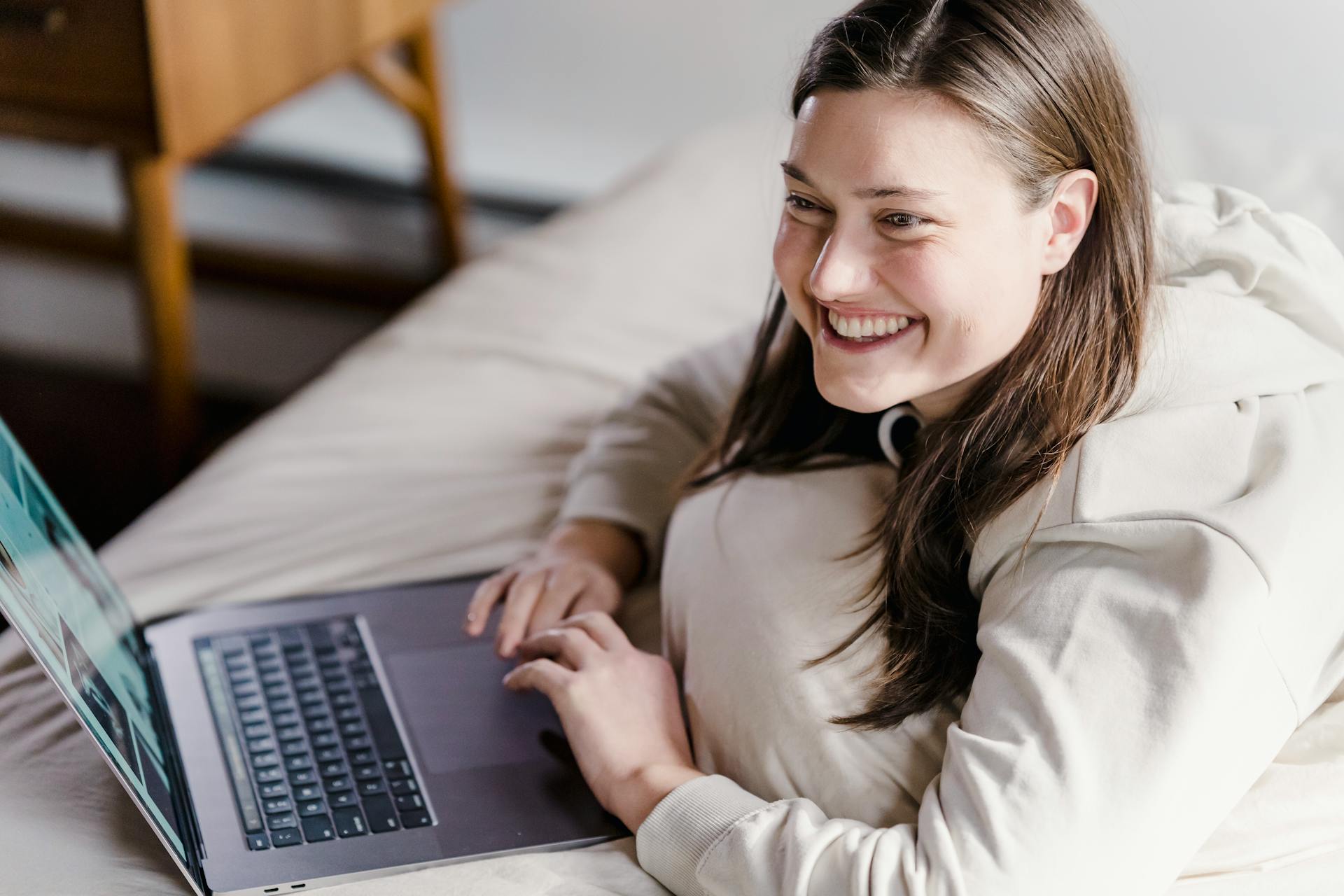 Happy female freelancer smiling and looking away while surfing modern netbook on soft bed in bedroom during remote work at home