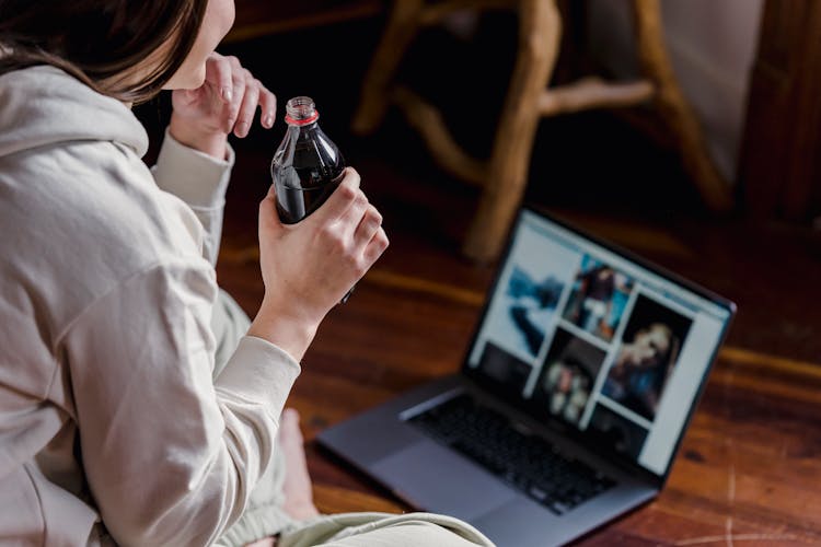 Crop Woman With Bottle Of Soda Sitting On Floor With Laptop