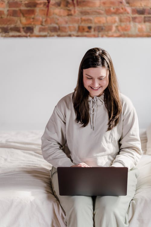 Smiling female freelancer typing on netbook while sitting on comfortable white bed near brick wall in bedroom during remote work