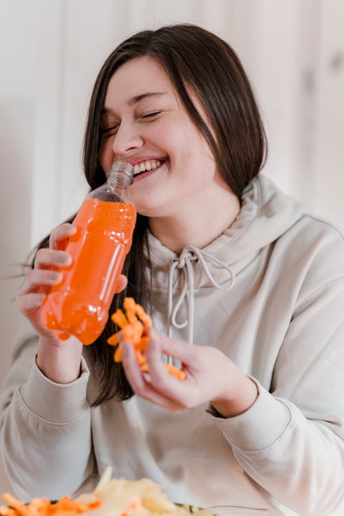 Glad young female in hoodie smiling and eating tasty fried crusty snack and drinking lemonade