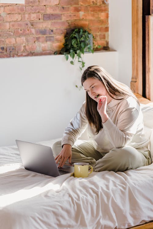 Free Young female resting on bed while using netbook and having snack in comfortable lounge Stock Photo