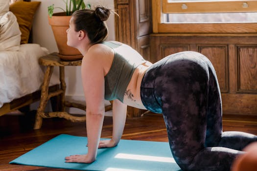 A woman practicing yoga indoors, performing a stretching pose on a blue mat by a window.